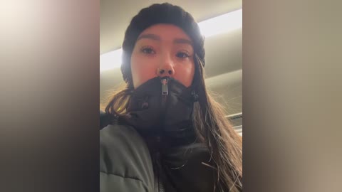 Media: A video shows a young woman with long, dark hair, wearing a black beanie and winter coat, with a black face mask over her mouth, taken indoors with fluorescent lighting.