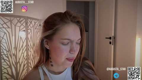 Media: Video of a young woman with light skin, long brown hair, and a delicate facial structure, wearing a white top, standing indoors next to a modern, geometric decorative screen.