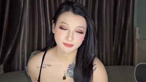 Media: Video of an Asian woman with long black hair, wearing a black spaghetti strap top, tattoos on her shoulders, sitting with a serene expression, against dark brown drapes.