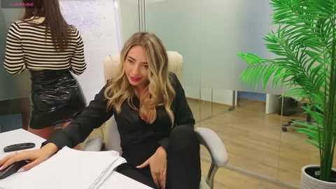 Media: Video of a blonde woman in a black blazer and skirt sitting at a desk in an office, while a brunette woman in a striped top and skirt stands behind her.