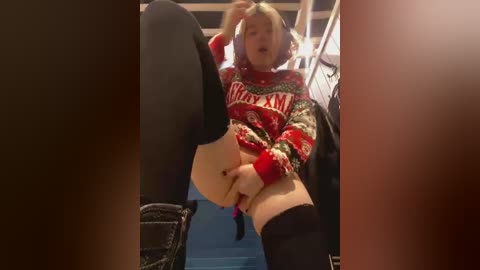 Media: Video of a blonde woman in a Santa sweater, black thigh-high stockings, and black boots, sitting provocatively with legs spread, revealing her vulva, in a dimly lit room.