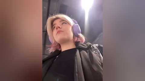 Media: Video of a young woman with light skin, short pink hair, and a purple beanie, wearing a black shirt and a grey jacket. The background is dimly lit with a bright light source at the top center.