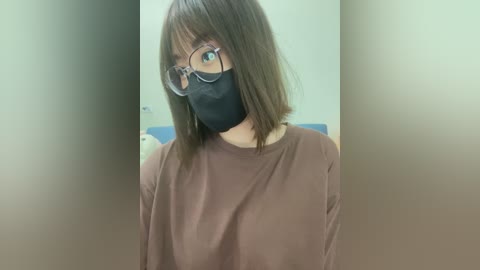 Media: Video of a young Asian woman with shoulder-length brown hair and glasses, wearing a black face mask and brown shirt. Background is blurred, possibly a room with a teddy bear.