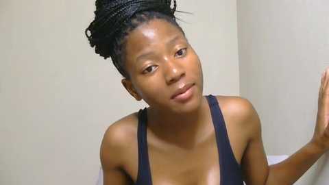 Media: Video of a young Black woman with braided hair, wearing a black tank top, standing indoors against a plain beige wall. She has a neutral expression and is raising her right hand.