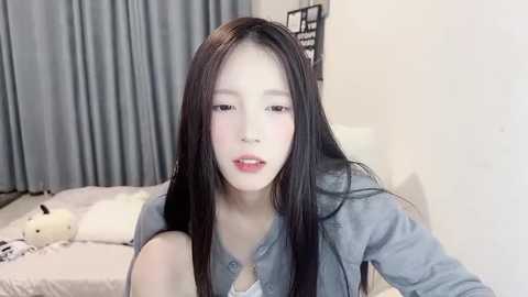 Media: A video of an East Asian woman with long black hair, fair skin, and light makeup, wearing a denim jacket, standing in a minimalist bedroom with white walls and blue curtains.