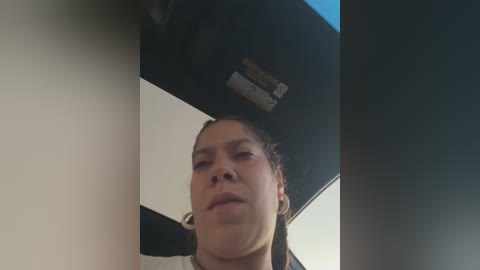 Media: Video of a young woman with light brown skin and dark brown hair tied back, looking down from a car's roof. She wears a white t-shirt. The background shows a blue sky and a black car roof.