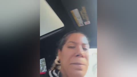 Media: A blurry video of a middle-aged woman with light skin, dark hair tied back, wearing large hoop earrings and a striped shirt, sitting in a car with a black interior.