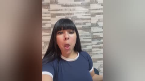 Media: Video of a young woman with long black hair and bangs, wearing a navy blue t-shirt, mouth open in surprise or shock, standing against a textured, monochromatic wall.