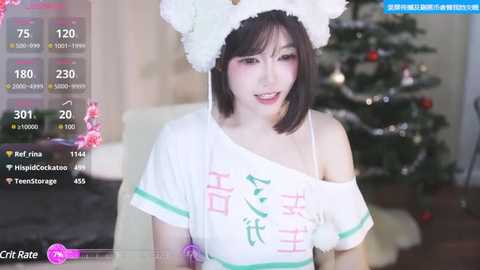 Media: Video of a young Asian woman with straight black hair and light skin, wearing a white off-shoulder crop top with Chinese characters and a white furry hat, sitting in front of a decorated Christmas tree.