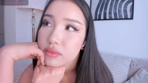 Media: Video of a young Asian woman with long black hair, light skin, and full makeup, leaning on her hand, looking thoughtful, in a modern, minimalist bedroom with a gray bedspread and abstract artwork.