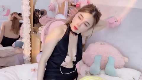 Media: A video of a slender, fair-skinned woman with long brown hair, wearing a black dress, lying on a bed with plush toys, in a pink-themed room.