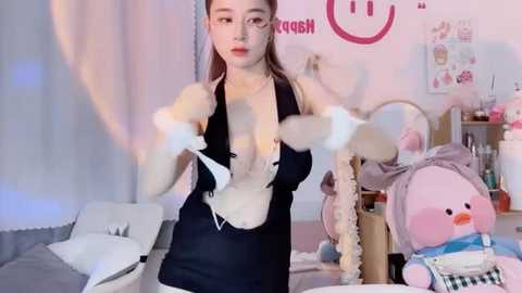 Media: A video of an East Asian woman in a revealing black maid outfit, with fluffy white gloves, in a colorful, cluttered bedroom with stuffed animals and posters.