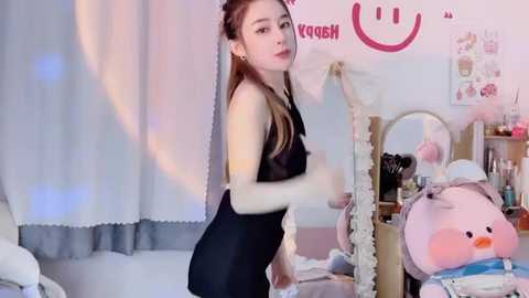 Video of a slim, fair-skinned woman with long, light brown hair, wearing a black sleeveless top and tight black pants, posing in a colorful, kawaii-themed room with plush toys and pastel decor.