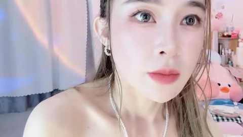 Media: Video of a young, fair-skinned woman with long, wet, light brown hair, wearing minimal makeup, including a silver nose ring and hoop earrings. She has a soft, natural look, with a subtle pink lipstick. The background features a pastel-colored room with a plush pink bunny.