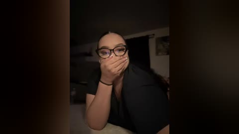 Media: A video of a young woman with light skin, wearing glasses and a black shirt, covering her mouth with her hands, in a dimly lit room.