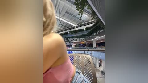 Media: Video of a blonde woman in a pink strapless top, seated indoors. The background shows a modern, glass-enclosed mall with greenery, shops, and people.