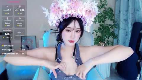 Media: A video of an East Asian woman with long black hair, wearing a floral headpiece and a revealing gray halter top, standing in a modern living room with blue lighting.