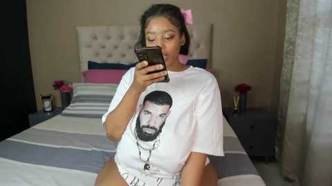 Media: Video of a young woman with medium skin tone and dark hair, wearing a white t-shirt with Drake's face, taking a selfie in a bedroom with a striped bedspread, pink flowers, and a tufted headboard.