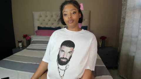 Media: Video of a young Black woman with short, dark hair adorned with a pink flower, wearing a white T-shirt with a graphic of a man's face, sitting on a bed in a modern bedroom.