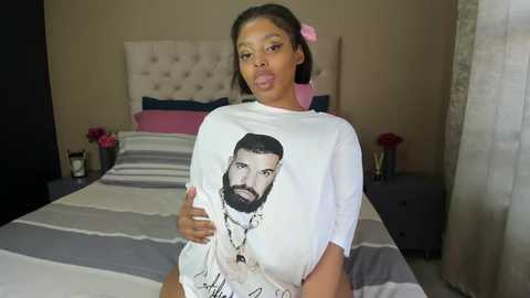 Media: Video of a Black woman with medium skin tone and short, straight hair, wearing a white T-shirt with a graphic of a man, in a modern bedroom with a striped bedspread and pink flowers.