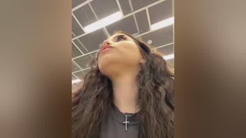 Video of a woman with long, dark, wavy hair, wearing a black top, and a silver cross necklace, looking upward against a grid ceiling with fluorescent lights.