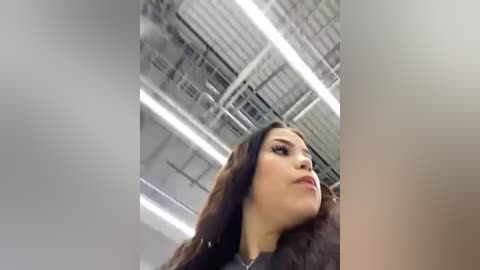 Video of a young woman with long brown hair, wearing a black top, looking upward in a modern, industrial setting with white and gray metal beams and fluorescent lighting.