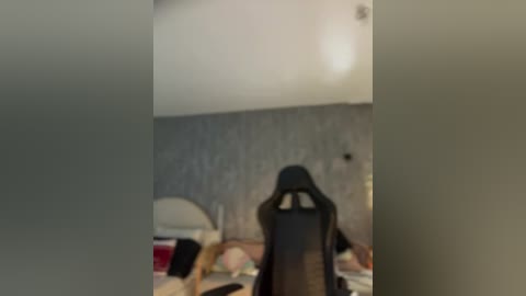 Media: Blurry video of a person in a gaming chair facing a bed with a white headboard and a grey wall, with a faint outline of a person in the background.