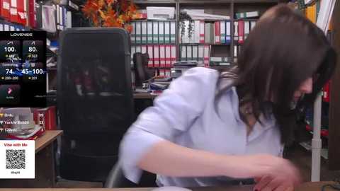 Media: Video of a woman with long brown hair, wearing a white shirt, working intently at a cluttered desk in a library-like office.