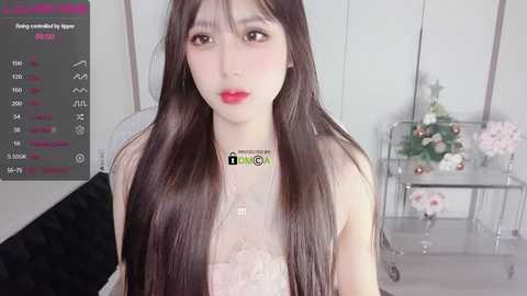 Media: Video of a young Asian woman with long, straight black hair and fair skin, wearing a delicate pink lace top, standing indoors in front of a floral arrangement and a metal cart.