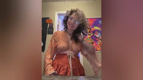 Media: Video of a middle-aged woman with voluminous curly hair and glasses, wearing an orange floral dress and a black apron, standing in a colorful, decorated room with psychedelic wall art.