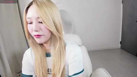 Media: Video of a young, fair-skinned woman with long blonde hair wearing a white T-shirt, sitting on a white chair in a minimalist, white-walled room with gray floor.