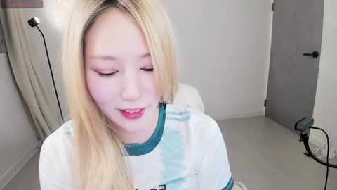 Media: Video of a young, fair-skinned Asian woman with long, straight blonde hair, wearing a white T-shirt with teal accents. She has a calm expression and is sitting in a modern, minimalist room with white walls and a grey door.