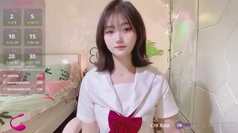 Media: Video of a young Asian woman with straight black hair and fair skin, wearing a white blouse with a red bow, sitting on a bed in a cozy room with a pink and green floral quilt.