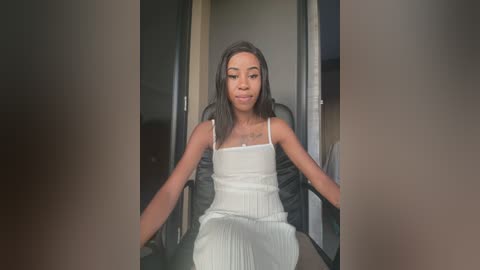 Media: Video of a young Black woman with straightened hair, wearing a white spaghetti-strap dress, sitting in a black office chair in a dimly lit hallway.