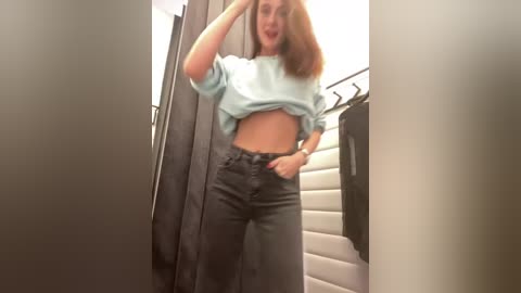 Media: Video of a young woman with light skin and shoulder-length auburn hair, wearing a light blue off-shoulder top and high-waisted black jeans, standing in a dressing room with grey curtains and a white wall.