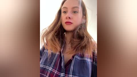 Media: Video of a Caucasian woman with fair skin and long, straight, reddish-brown hair. She wears a blue plaid shirt and has a neutral expression. Background is blurred, focusing on her.