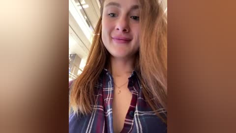 Media: Video of a young woman with light skin, straight brown hair, and a slight smile, wearing a red and blue plaid shirt, standing in a subway car with blurred background.