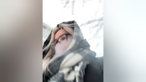 Media: Video of a young woman with fair skin, wearing glasses, a gray and white striped scarf, and a black jacket, partially obscured by snowfall, standing in a snowy, wintry background.