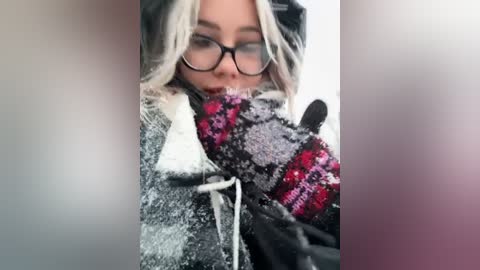 Media: A close-up video of a woman with long, platinum blonde hair, wearing black-rimmed glasses, a grey hoodie, and a colorful, patterned scarf, slightly blurred, with a soft, muted background.