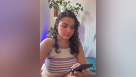 Media: Video of a young woman with curly black hair, wearing a striped sleeveless top, sitting on a bed, holding a smartphone, surrounded by indoor plants, and a blue blanket.