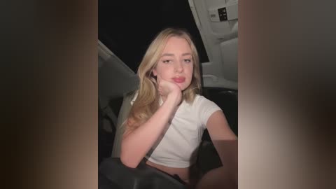 Media: Video of a young Caucasian woman with long blonde hair, wearing a white crop top, sitting in a car at night, looking contemplative.