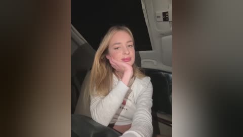 Media: Video of a young Caucasian woman with long blonde hair, seated in a car at night, resting her chin on her hand, wearing a white sweater, and looking pensive.