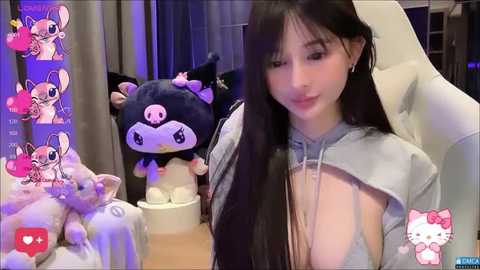 Media: A young East Asian woman with long black hair, wearing a gray hoodie, sits in a modern room filled with plush toys, including a large black cat and a Hello Kitty.