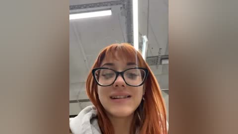 Media: Video of a young woman with long, straight red hair and black-framed glasses, smiling, set against a modern industrial background with exposed pipes and metal structures.