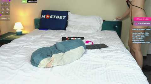 Media: Video of a woman wearing a black bra and panties standing on a bed, with a plush shark pillow and a remote control on the white sheets.