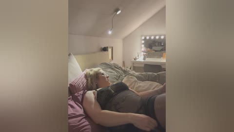 Media: A video of a woman with blonde hair, lying on a messy bed, wearing a black top and shorts, in a dimly lit, cluttered bedroom.