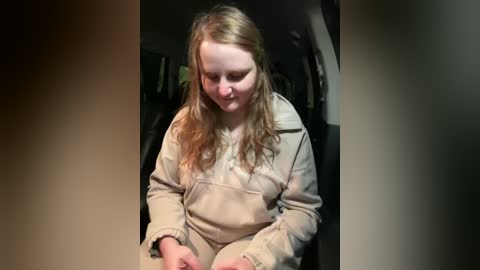 Media: Video of a young, fair-skinned girl with long, wavy blonde hair, wearing a beige sweatshirt and pants, sitting on a sofa in a dimly lit room.