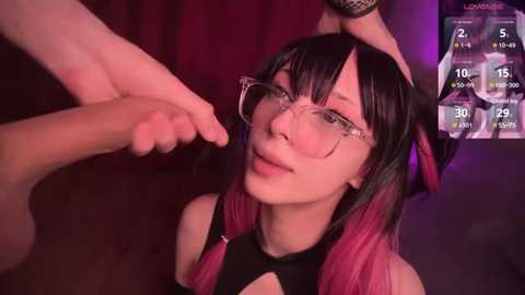 Video of a young, fair-skinned woman with long black hair and pink streaks, wearing glasses, being touched on the head by a man's hand. Background features a dark red curtain and a digital screen displaying weather information.