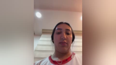 Media: A video of a young woman with light skin and dark hair, wearing a white shirt with red trim, standing in a modern, brightly lit hallway with white walls and a staircase in the background.