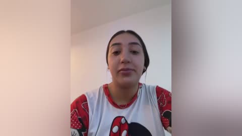 Media: Video of a Latina woman with medium skin tone, long black hair in a bun, wearing a red and white Mickey Mouse shirt, standing in a simple, light-colored room.